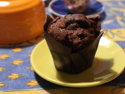 Muffin al cioccolato fondente servito su un piattino giallo, dettaglio di un dolce soffice in pirottino di carta scura su tavola colorata per colazione o merenda