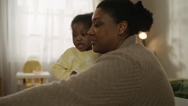 Side view handheld shot of caring young African American mother interacting with adorable toddler daughter while sitting at table at home in modest apartment and giving her cookie