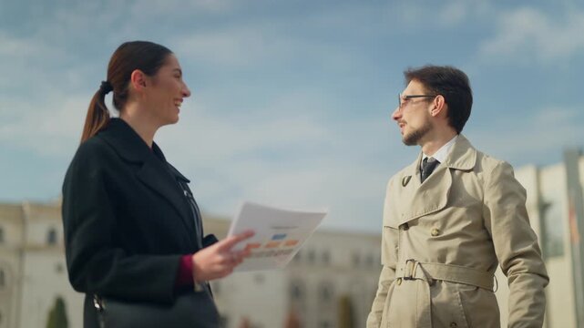 Two business professionals hold an outdoor meeting, discussing documents with charts and graphs. They engage in a focused conversation in an urban environment, exchanging ideas.