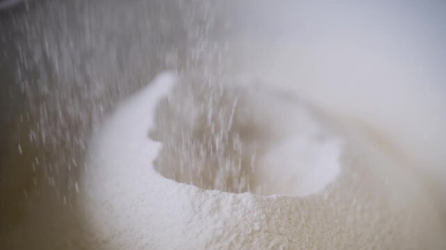White flour sifts down into a large mixing bowl in slow motion. The fine particles create a delicate dust cloud as they pile up to form the base for dough or pastry preparation.