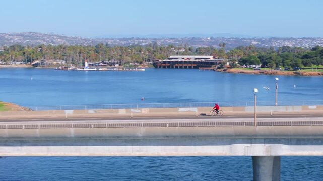 Aerial view of Mission Bay Park in San Diego California featuring concrete bridge with cars and cyclist in red shirt crossing over blue water.