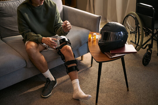 Young adult man with disability taking pain medication while sitting with leg brace after motorcycle road accident. Useful for rehabilitation, injury recovery, insurance or healthcare campaigns