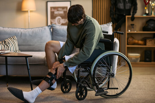 Young adult Caucasian man with disability sitting in wheelchair, adjusting leg brace during home recovery. Useful for rehabilitation, mobility support, healthcare marketing campaigns