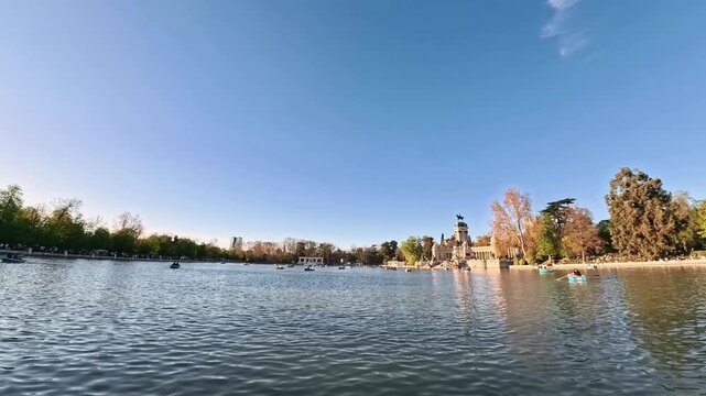 Wide shot of people rowing blue boats on the Estanque del Retiro with the Monument to Alfonso XII in the background during a sunny afternoon in Madrid, Spain.