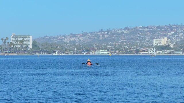 Person in orange life vest and straw hat kayaks across tranquil blue bay waters. Coastal cityscape with hillside homes, palm trees, hotels, and sailboats visible in hazy sunny conditions