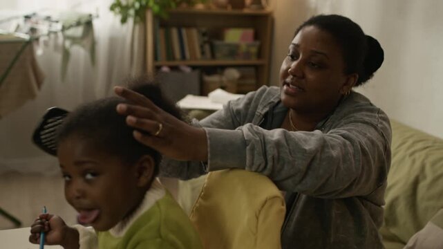 Handheld shot of caring young African American mother sitting on couch in modest apartment and combing hair of joyful toddler daughter sitting in high chair and willing to draw in notebook
