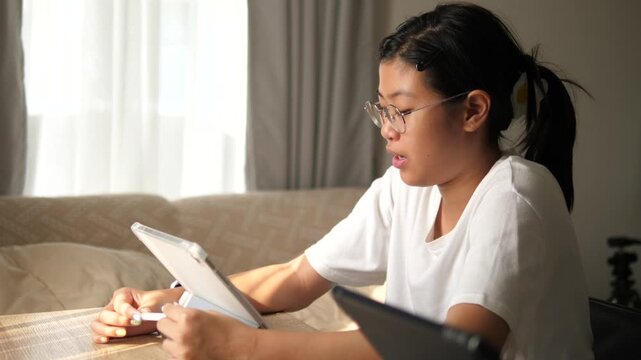 Focused young Asian girl wearing glasses using a digital tablet and stylus for online learning at home with warm sunlight background, education and technology concept