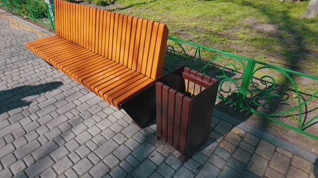 A bright orange wooden park bench sits empty next to a dark brown slatted trash bin on a sunny day. They rest on a grey paved walkway bordered by a green metal fence and fresh grass.