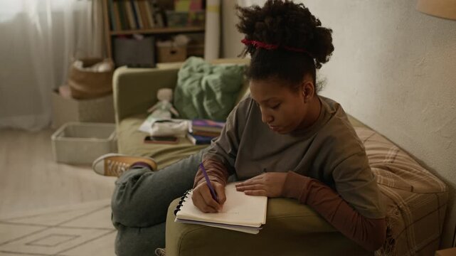 Zoom in shot of tired African American teenage girl writing in notebook while sitting on couch at home surrounded by educational materials and then closing eyes feeling tired of studying