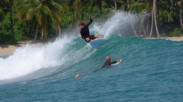 SLOW MOTION: Surfer makes a powerful top turn, sending a burst of white spray into the air above blue wave. Palm trees and tropical shoreline frame the action at Beng Bengs surf spot in Mentawais.