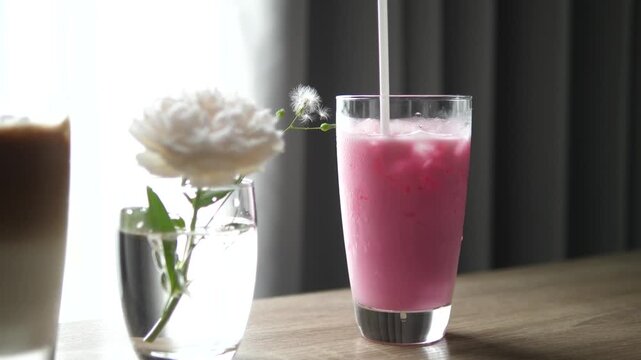 Close-up of refreshing iced pink milk (Nom Yen) in a glass on a wooden table with a white flower in a vase, sweet beverage and lifestyle concept