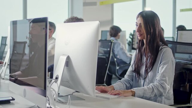 Young businesswoman working at desktop computer in a modern open plan office. Focused female employee sits at her workstation and handles daily tasks while colleagues work at nearby desks in the
