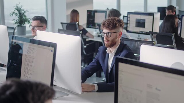 Professional call center team working at computers in a bright open plan office. Young male support agent in formal suit sits at workstation while colleagues work in the background, creating a