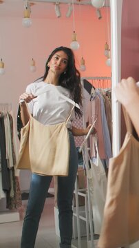 Stylish young woman choosing clothes and comparing outfit options while looking at her reflection in a mirror inside a modern fashion boutique. Female shopper holds hangers with garments and evaluates