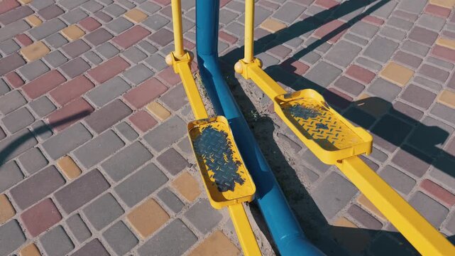 Yellow and blue foot pedals of an outdoor elliptical trainer stand empty in the sun. The painted metal shows signs of wear and rust on the diamond plate texture. Colorful paving stones surround it