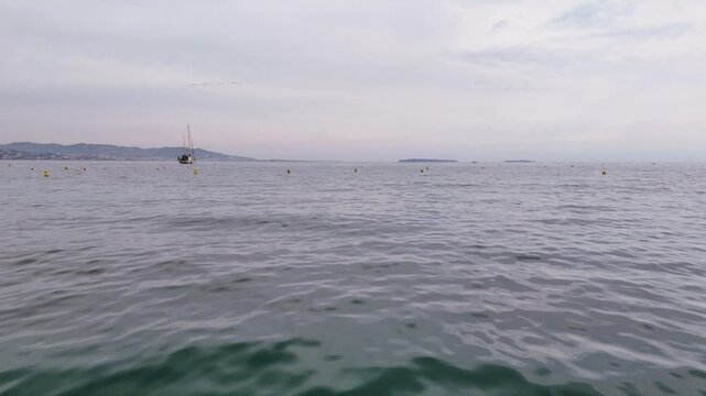 Yellow Buoys Marking Calm Coastal Waters, Small Moored Sailboat, Gentle Swell Near Shoreline, Misty Horizon And Nautical Atmosphere Ideal For Navigation And Harbor Approach Footage
