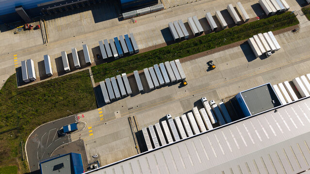 Aerial view of Magna Park logistics hub featuring rows of white semi-trailers parked outside large industrial warehouses under bright sunlight in Magna Park, England, United Kingdom.