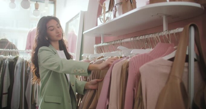 Stylish young woman browsing clothing rack and choosing garments on hangers in a modern fashion boutique. Female shopper in a pastel blazer examines apparel in a soft pink retail interior surrounded