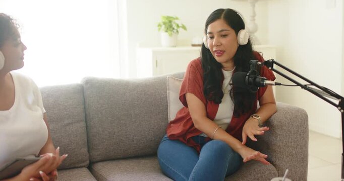 Turning right-side host wearing white headset speaking into mic on gray sofa, recording podcast