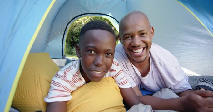 African American father and son smiling, leaning toward camera inside blue dome tent with metal mug
