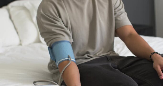 Man in 20s sitting on bed adjusting blue blood pressure cuff while checking white monitor smiling