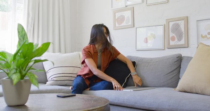 Rummaging woman in rust top, jeans searching dark pillow on sofa at home, with tablet, smartphone