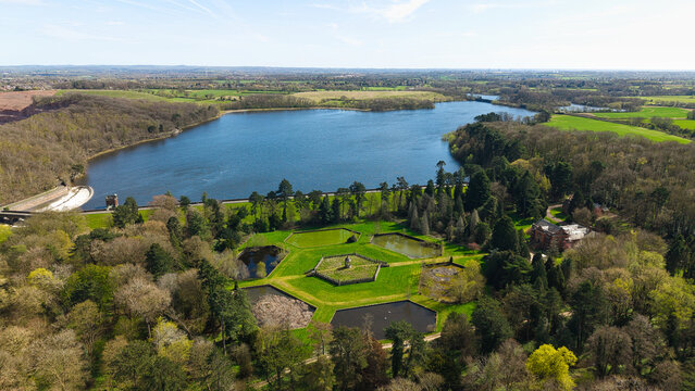 Aerial view of Swithland Reservoir featuring hexagonal ponds, a dam wall, and dense green woodland under a clear blue sky in Leicestershire, England, United Kingdom.
