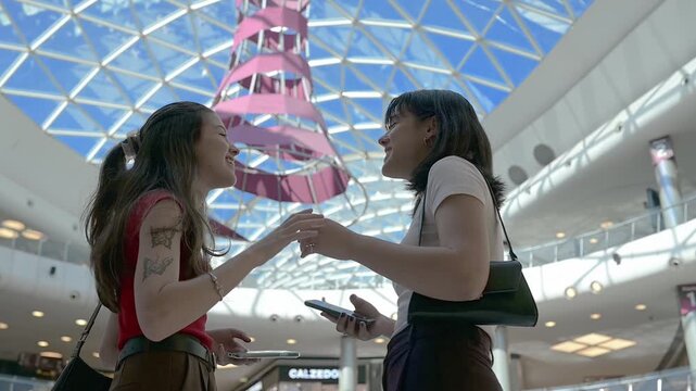 Two young women enjoying leisure time and gesturing as they talk and laugh together while holding their smartphones inside Marineda City, Galicia, Spain. Lifestyle and friendship concept in a modern c