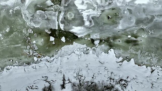 Aerial top down video of frozen Baltic Sea at T&uuml;risalu, Estonia, textured ice surface with cracks, snow patches and greenish water beneath. Cold winter weather, abstract natural pattern