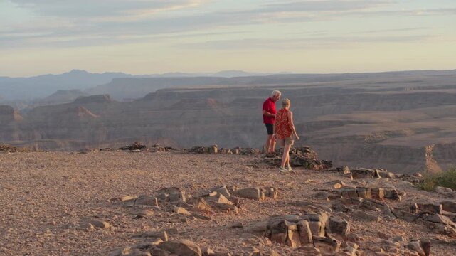 Aged European couple walk to cliff edge, take photographs of Fish River canyon in sunset light