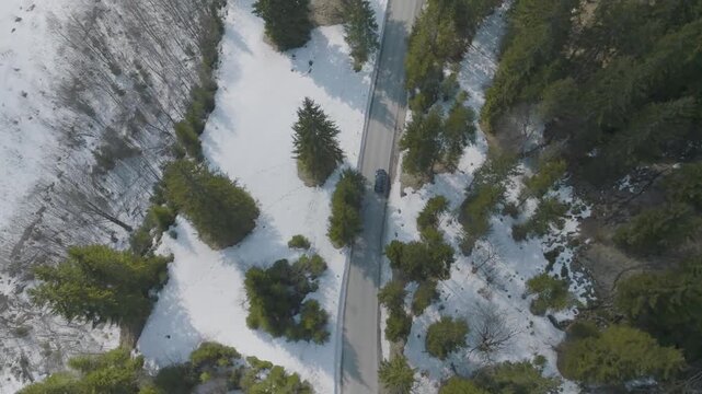 Aerial view of a car driving on a road cutting through a snowy landscape and dense forest, Lenggries, Bavaria, Germany.