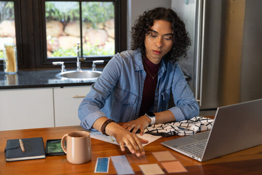 Non-binary adult in denim shirt arranging color swatches at home kitchen table consulting laptop