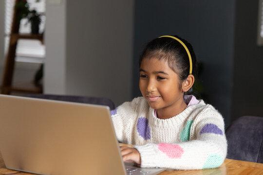 Indian child girl sitting at wooden table using silver laptop near window, wearing yellow headband