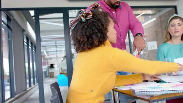 Woman in yellow top reaching docs, team pointing, sorting design mockups on glass table for review