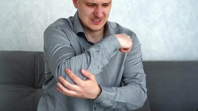 Young European man sitting in a bright room, holding his elbow and showing signs of pain after strain or overuse during physical activity or work.