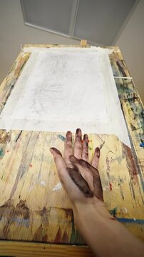 Charcoal-smeared artist hand poised before a taped blank drawing sheet on a paint-splattered wooden board, revealing the messy preparation stage of sketching in a studio