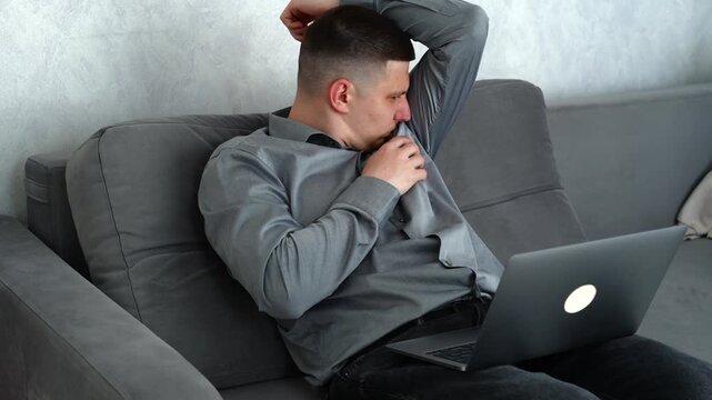 Man working remotely on a sofa in a warm room, feeling overheated and self-conscious due to sweating caused by hot weather while continuing work.