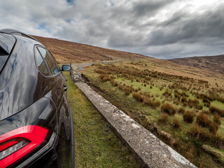Fototapeta premium Dark SUV parked beside winding rural road. Overcast sky looms above rolling green hills. Vehicle reflects misty, moody atmosphere. Path stretches into distance, inviting exploration Travel concept