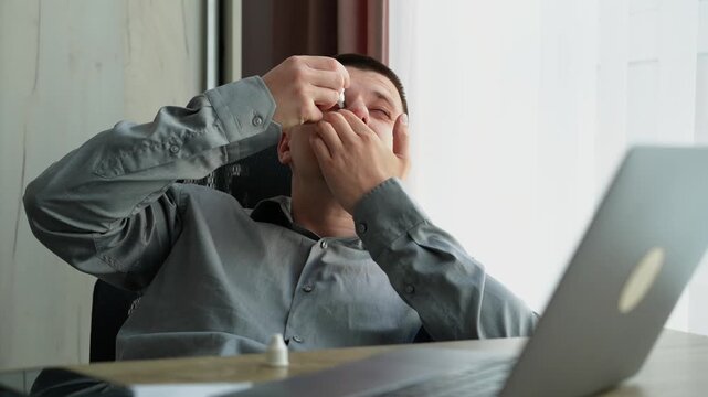 Close-up of a young male applying eye drops while sitting at a workplace desk, treating eye irritation caused by prolonged computer use.