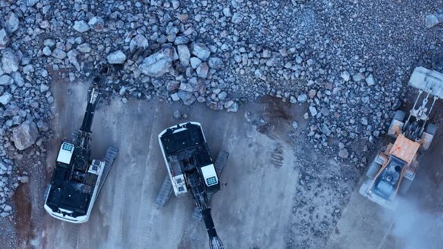 An aerial view captured by a drone of excavators and loaders operating at a quarry. An industrial mining operation and the rock-breaking process involving heavy machinery.