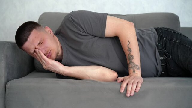 Brunette man lying on a gray sofa, waking up from a short rest and rubbing his face, expressing sleepiness and relaxation.