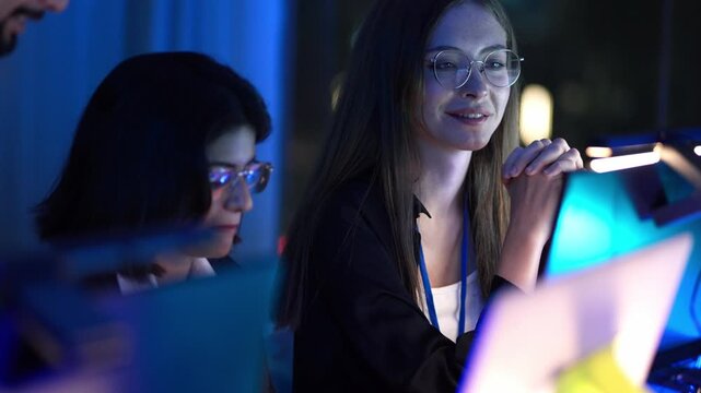 Smiling female software developers working on AI technology in dark office. Professional programmers collaborating on coding project with blue light reflection on screens for IT corporate success