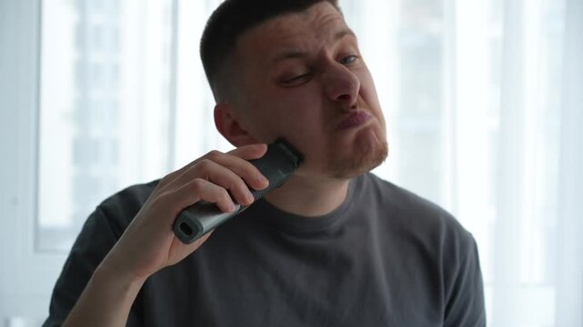 Young man carefully trimming facial hair with an electric razor in a well-lit room, maintaining a clean and styled beard. Personal care routine.
