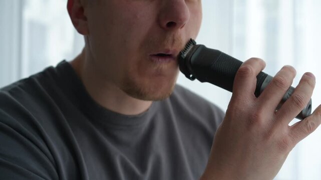 Young brunette man trimming his beard and mustache with an electric trimmer in a bright room, shaping facial hair as part of his daily grooming routine before going out.