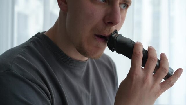 Brunette man grooming his beard and mustache with a trimmer in a bright indoor setting, focusing on precision and appearance before an important event.