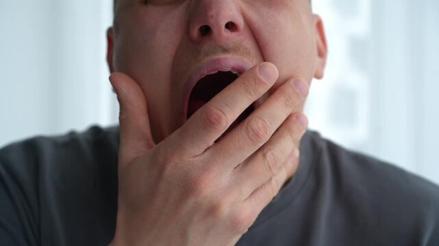 Close-up of a young man yawning in a bright room, showing signs of sleepiness in the morning. Concept of fatigue and waking up.