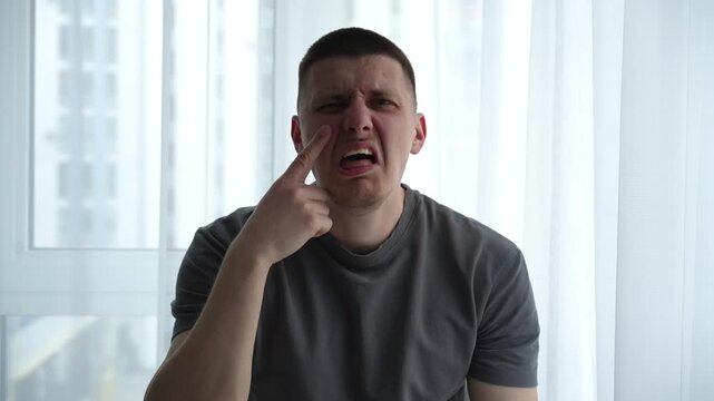 Man in a bright room acting out a fake crying motion with his hands on his face, showing a teasing and mocking expression as if imitating someone being overly emotional.