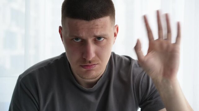 Close-up of a young male making a hand wave gesture that looks like a greeting, while his facial expression shows frustration and annoyance. Concept of conflicting emotions