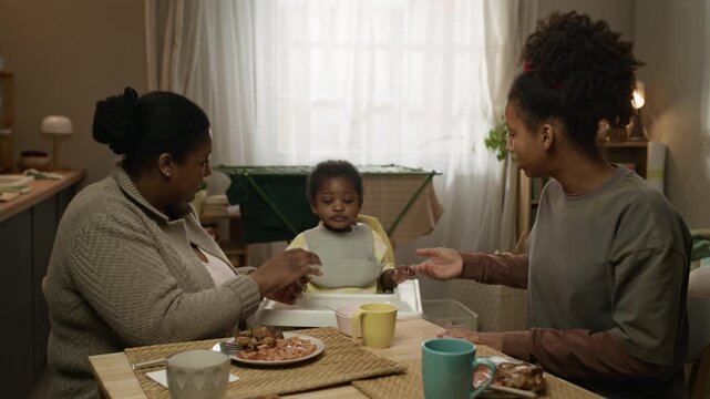 Medium shot of Black mother and teenage daughter sitting at table holding hands of toddler girl in high chair and praying together before sharing modest meal, living in small low income apartment