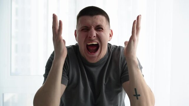 Young man shouting in frustration, showing anger and disappointment while holding his head in a bright room. Focus on emotional stress and feeling of lack of control.